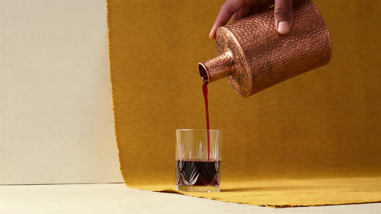 A hand tips a hammered copper bottle, pouring a thin oxblood stream into a cut-crystal glass on a mustard linen cloth against a cream paper wall