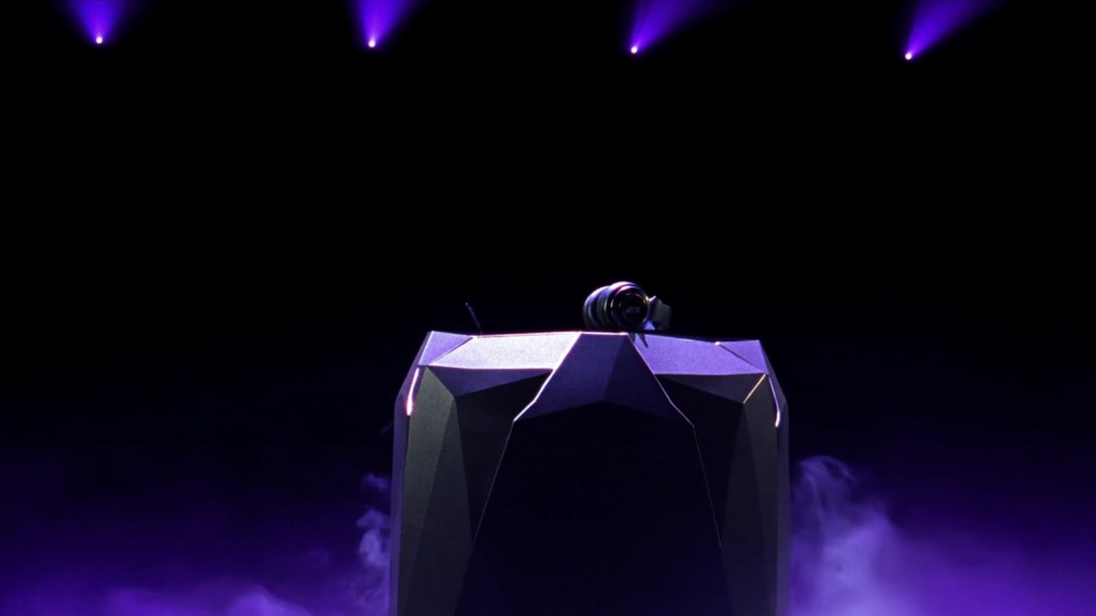 A black headset on a faceted chrome pedestal on a dark arena stage, violet beams cutting through low haze