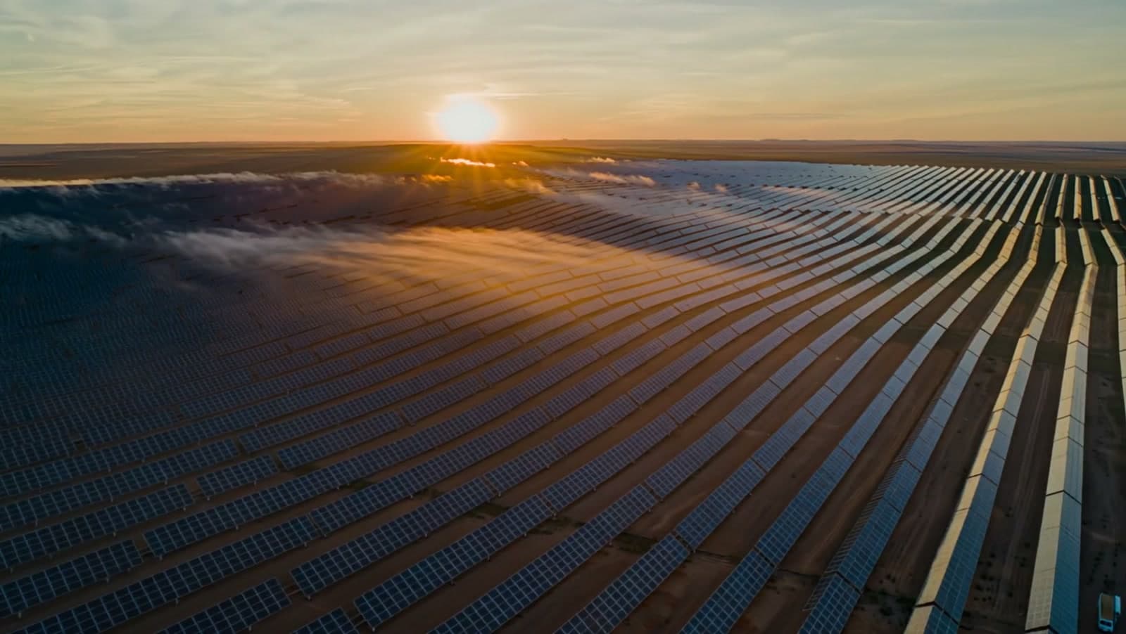 A vast solar farm at dawn seen from above, golden sun catching thousands of dark panel rows through morning mist