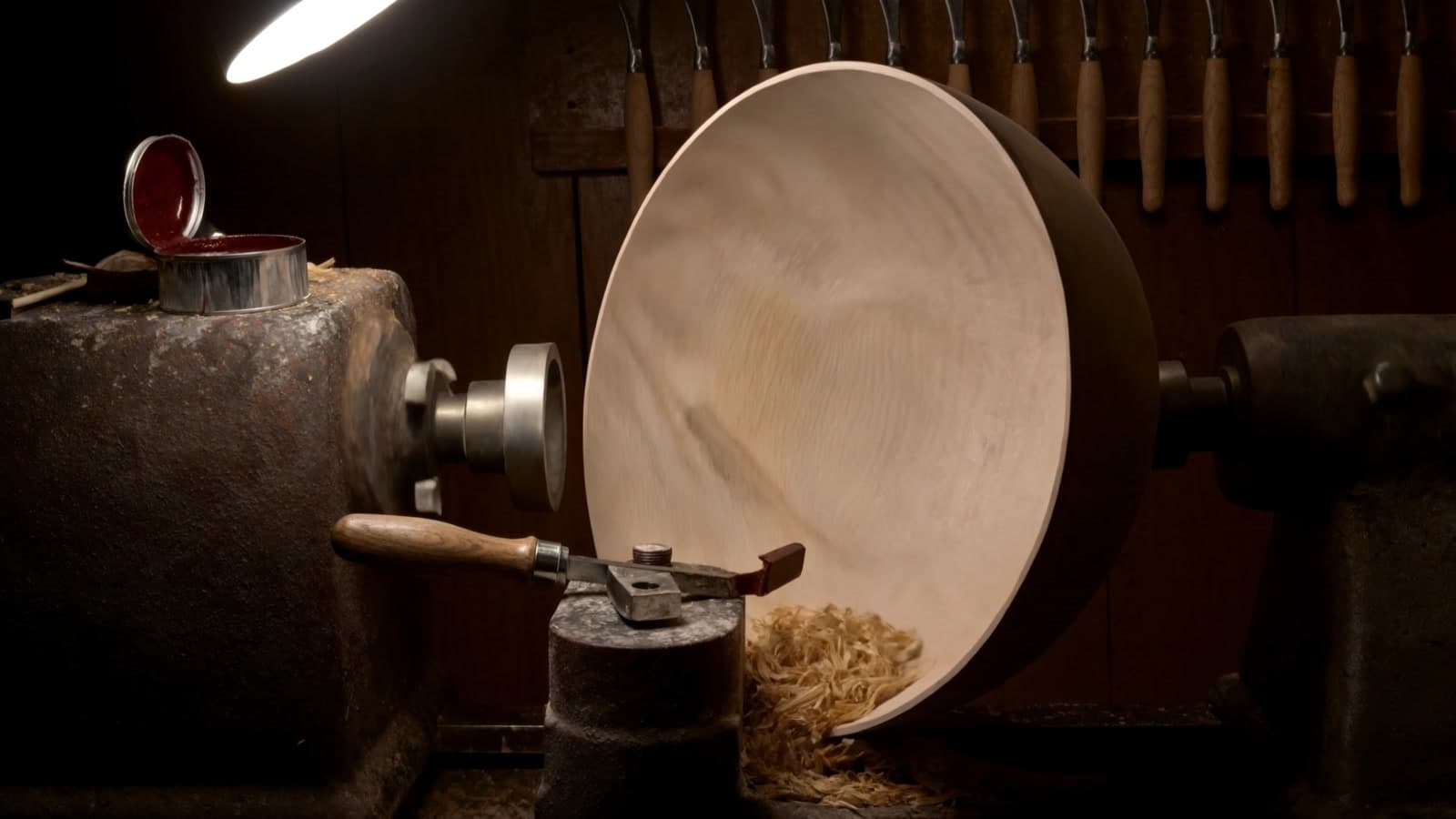 A pale turned bowl on a cast-iron lathe under a work lamp, a gouge on the tool rest, shavings below and an open tin of oxblood lacquer on the headstock