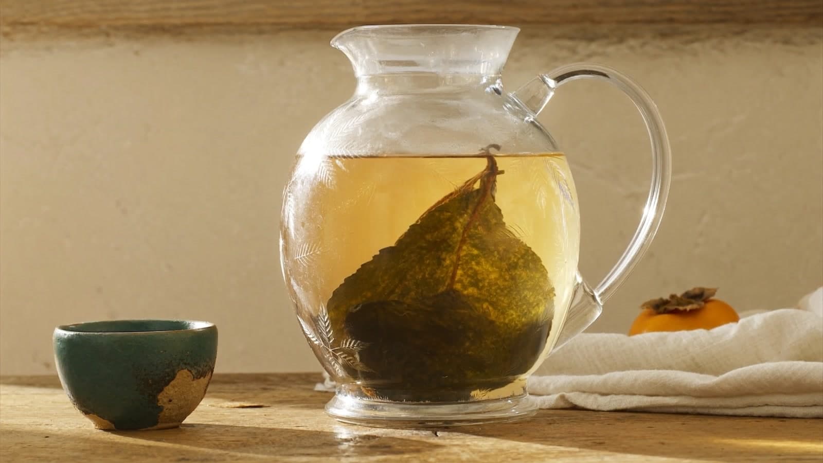 A clear glass jug of amber tea on an oak table in low sun, a dark whole tea leaf unfolding inside, a green glazed cup to the left and a persimmon on cream linen behind