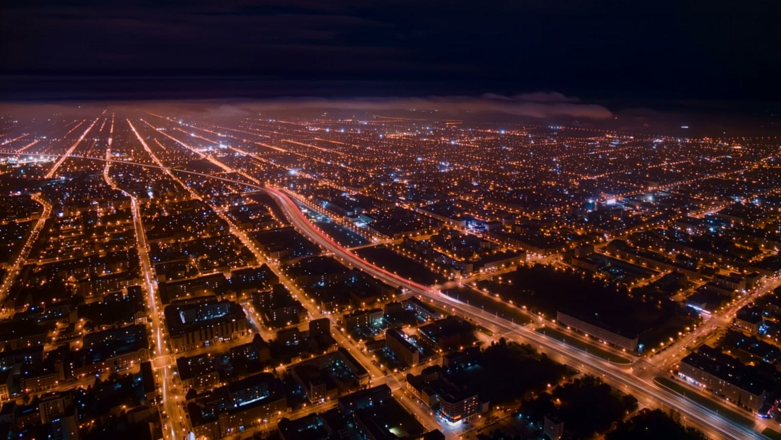 A vast city grid at night from high altitude, streets drawn in amber and periwinkle light