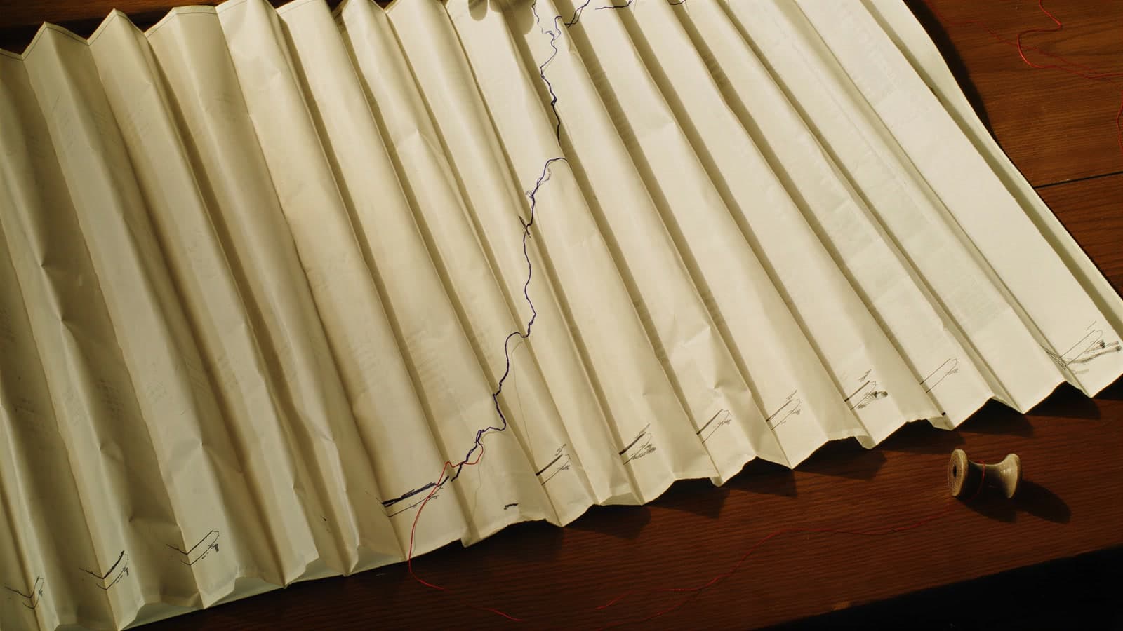 A sheet of cream paper folded into pleats on an oak desk in low sun, a blue ink line climbing across the folds, a loose red thread and a wooden spool on the wood