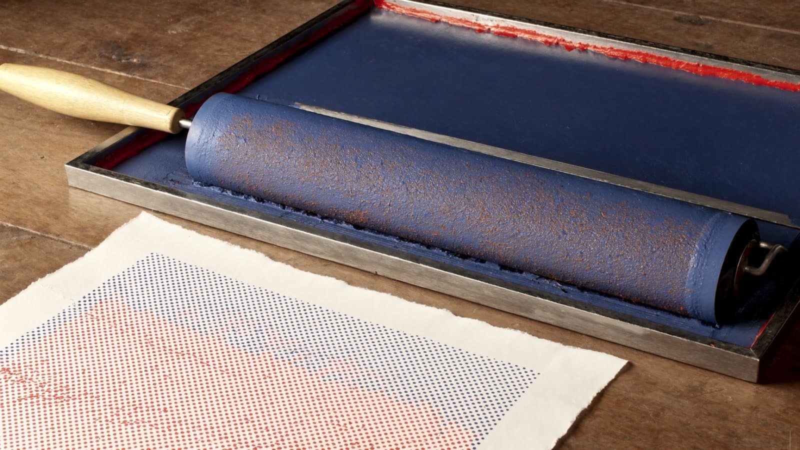 A brayer coated in ultramarine ink resting in a steel inking tray on a worn wooden bench, a ridge of vermilion ink along the tray's far edge and a deckled sheet of rag paper printed with blue and red halftone dots in front