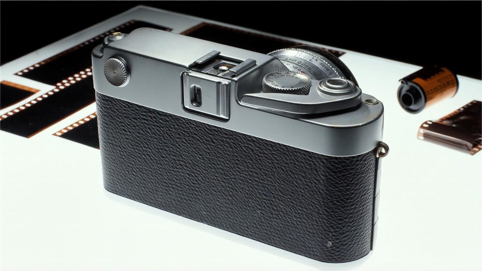 The back of a chrome and black rangefinder camera on a glowing white light table, black negative strips behind it and an orange film canister to the right
