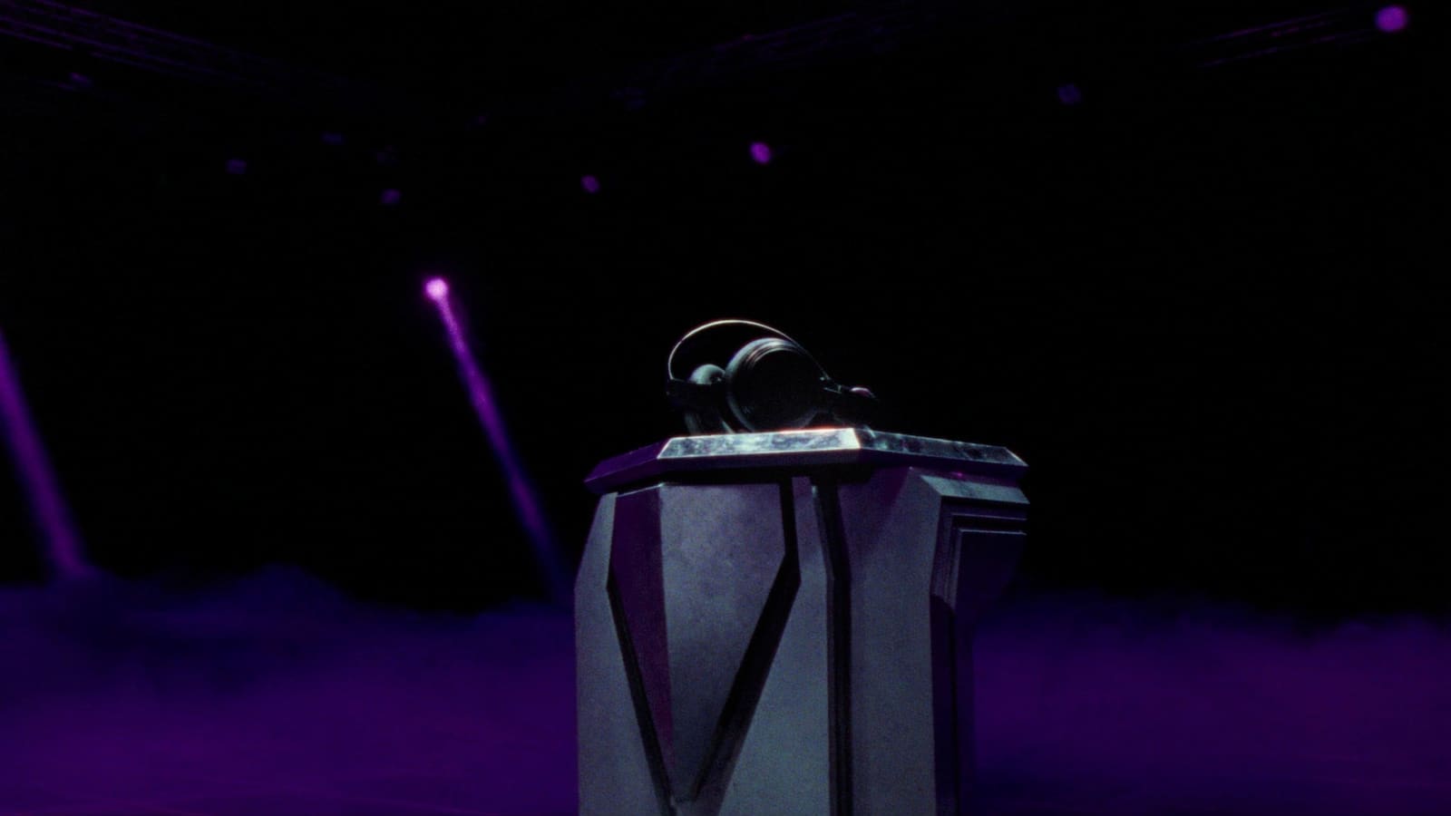 Reel frame: the black headset on its faceted pedestal, two violet beams cutting down through the haze