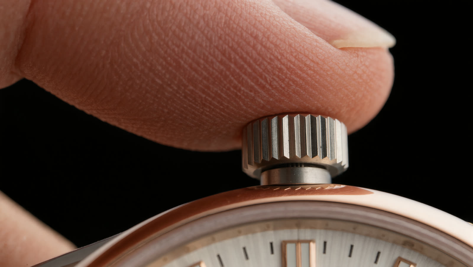 A fingertip resting on a fluted steel crown above a rose gold case and silvered dial