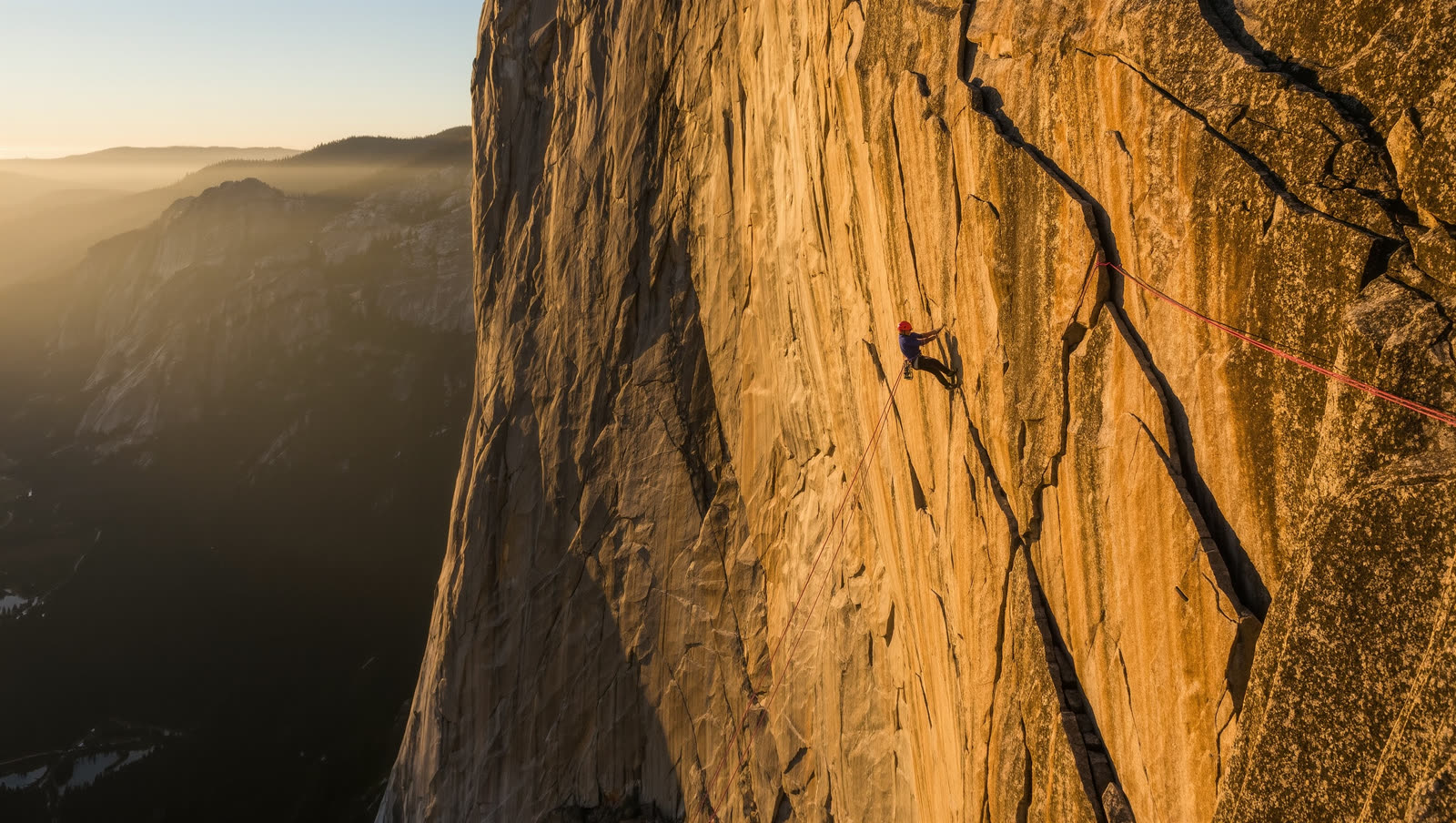 A climber on a red rope halfway up a sunlit granite wall above a hazy valley